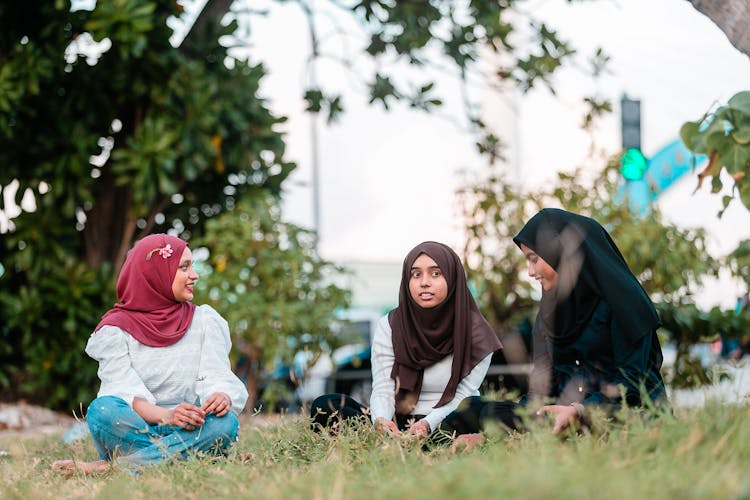 Happy Muslim Women Sitting On Green Lawn And Talking