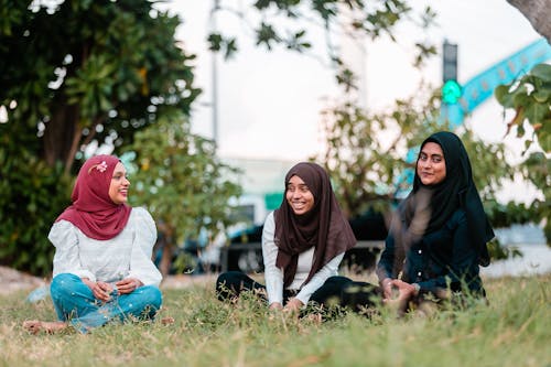 Free Full body joyful ethnic females in hijabs gathering together on grassy meadow in city park and chatting Stock Photo