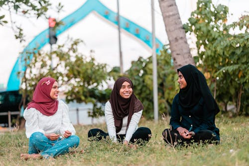 Full length cheerful young ethnic females in headscarves sitting on lush grassy park lawn and having conversation while looking at each other with toothy smile