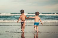 Back view of anonymous shirtless little brothers holding hands and walking on wet sandy beach towards waving ocean during summer holidays