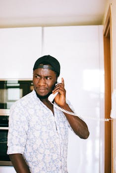 A serious man in casual attire using a landline phone indoors, appearing thoughtful.