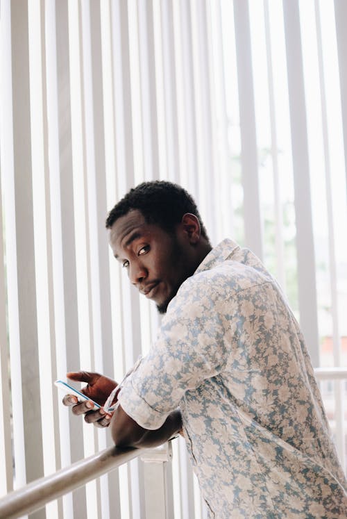 Serious young African American male in shirt with floral print looking at camera over shoulder while leaning to railing next to window decorated with blinds
