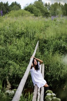 一位身穿白裙的女子坐在乡村桥上，四周郁郁葱葱的绿植环绕，构成一幅宁静的景象