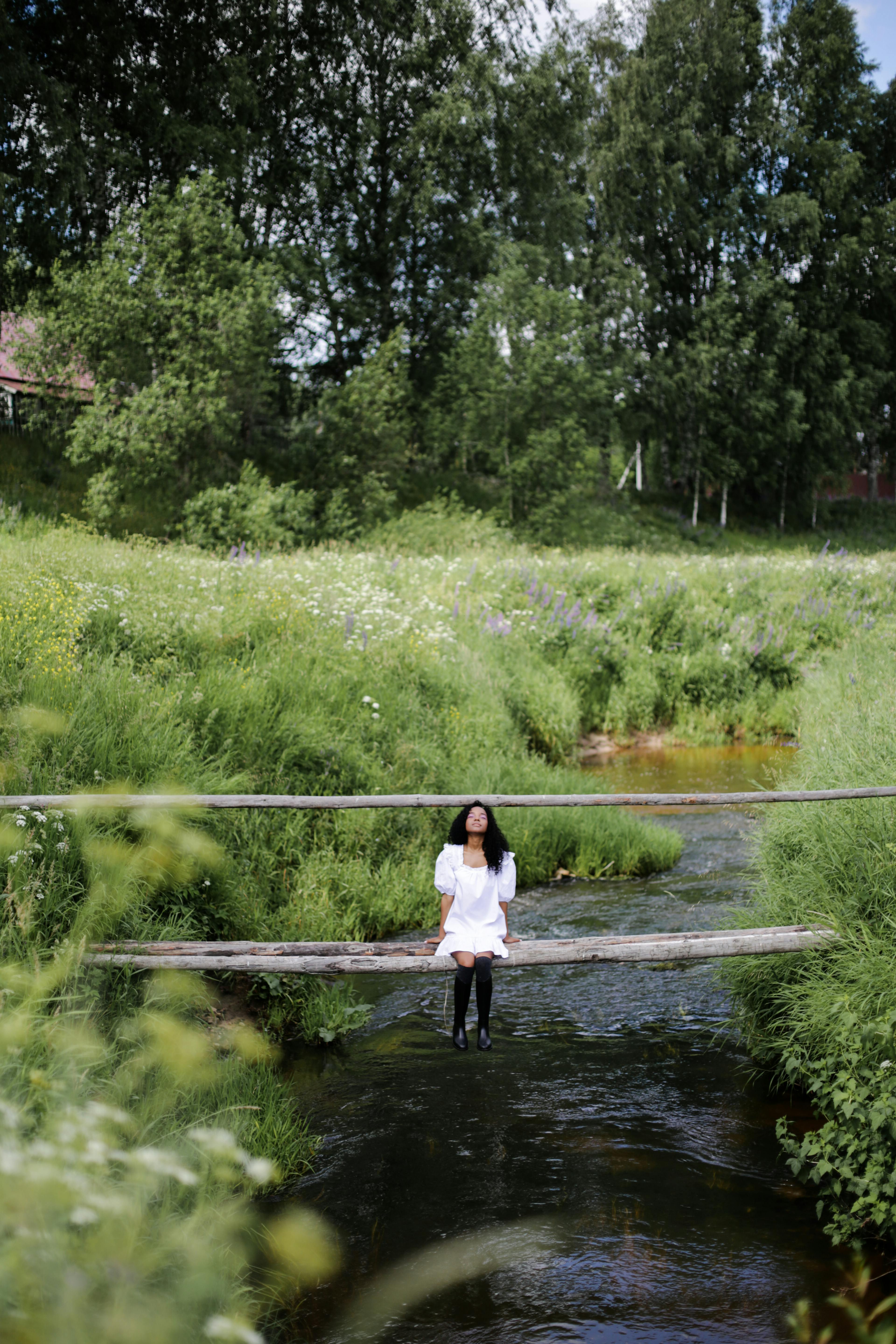 一名妇女坐在乡村桥上，桥下是郁郁葱葱的溪流，周围是绿色的乡村环境