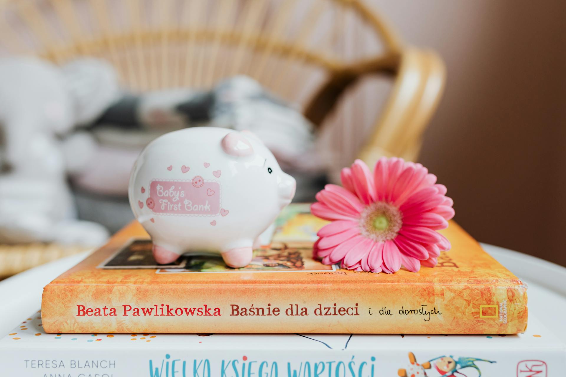 A piggy bank and pink gerbera flower rest on colorful children's books, suggesting childhood savings.