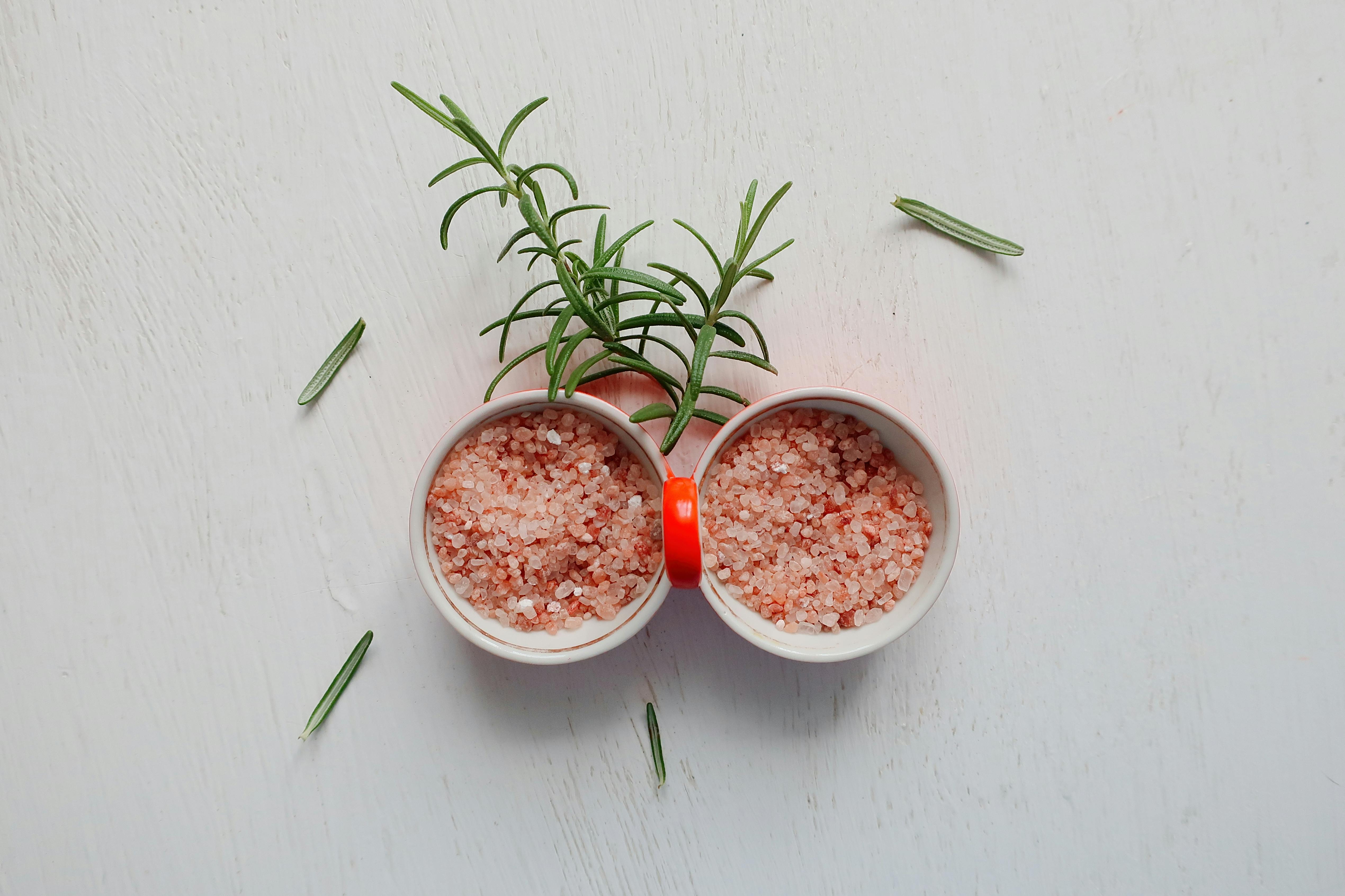 Top view of pink Himalayan salt and fresh rosemary in bowls on white table.