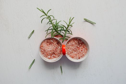 Top view of pink Himalayan salt and fresh rosemary in bowls on white table.