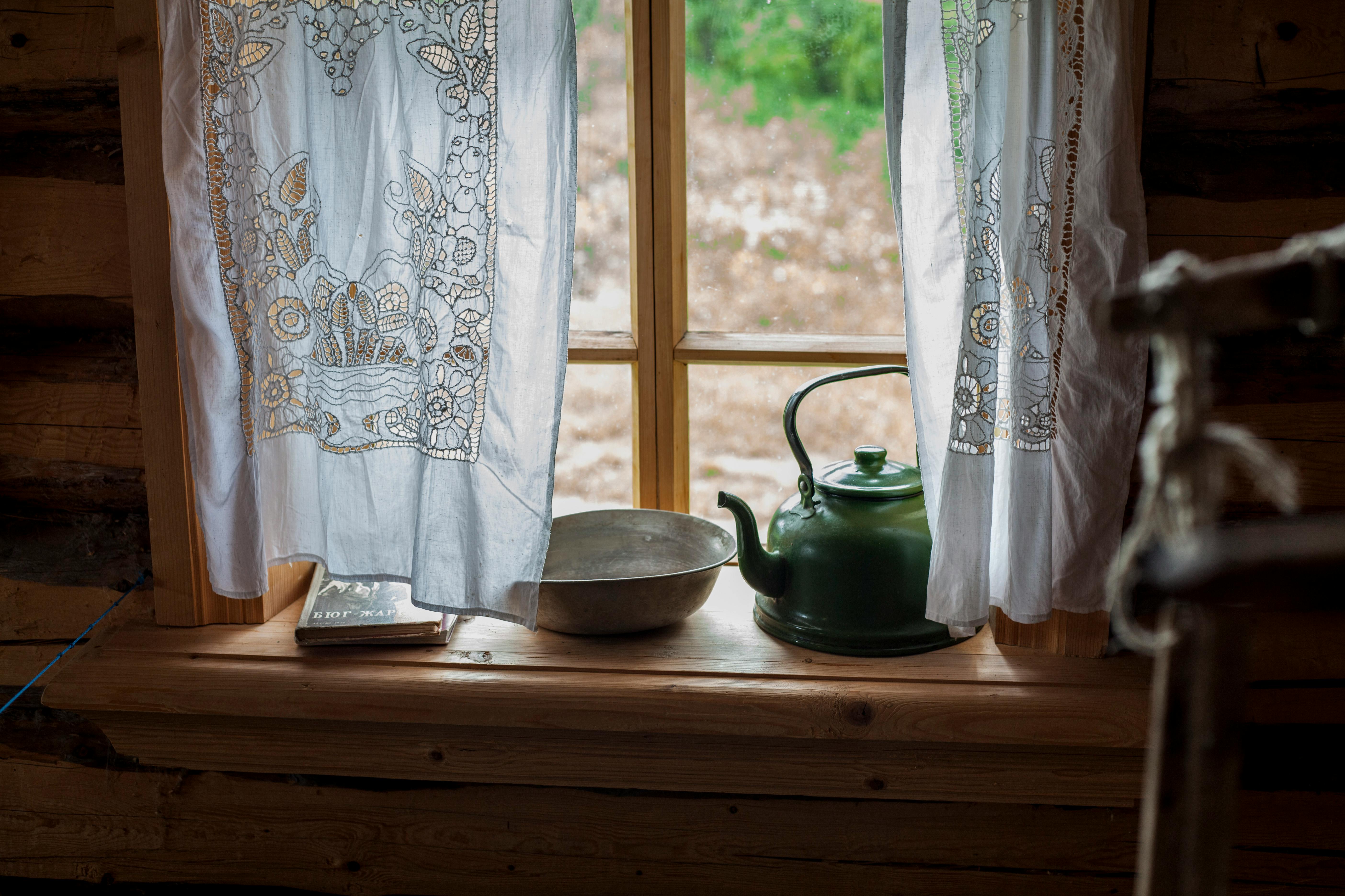 A cozy rustic windowsill with a green teapot and lace curtains overlooking the countryside.