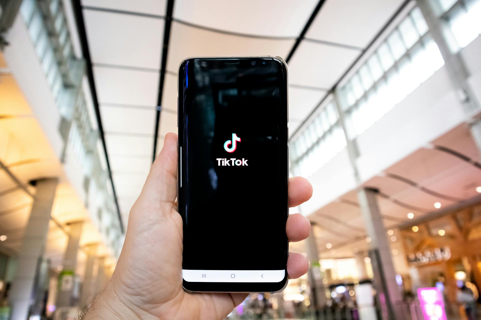 Close-up of a hand holding a smartphone displaying the TikTok logo in a public indoor setting.