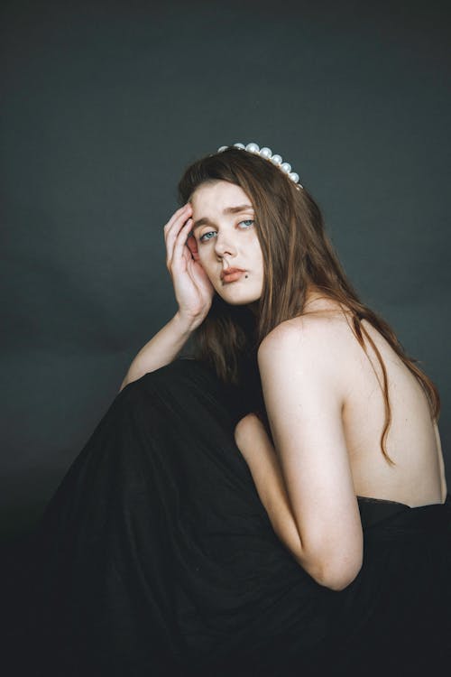 Unemotional young beautiful shirtless female with long straight hair decorated with tiara touching hair and looking at camera on gray background