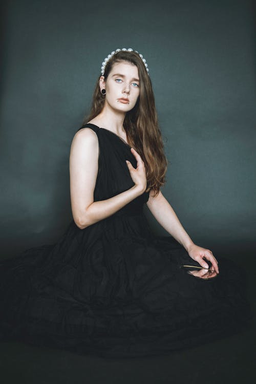 Pensive allure slender female in long black dress and tiara in hair touching chest and looking at camera in dark studio