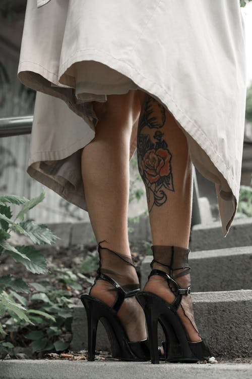 Unrecognizable stylish female in long coat and black high heels with tattoo of flower on leg on stairway in daytime