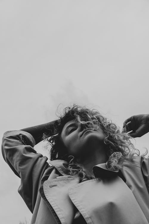 Black and white low angle of pensive young female in warm coat touching curly hair and looking ahead