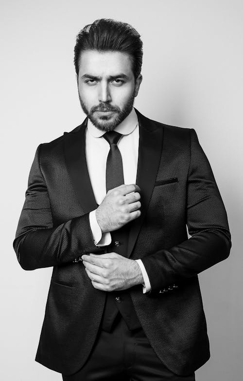 Black and white of bearded pensive strict male with trendy hairstyle in tie and formal outfit looking at camera