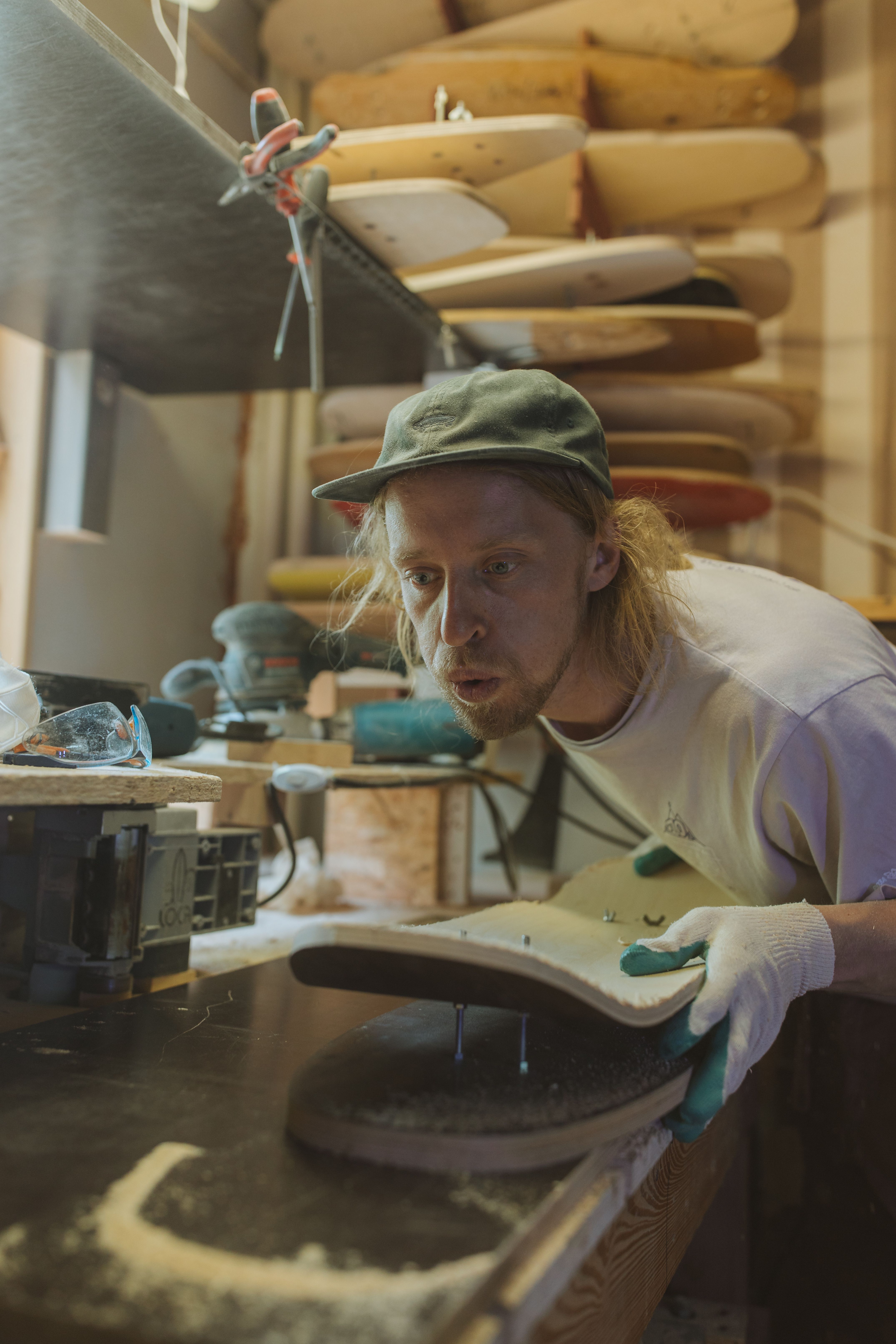 A focused craftsman shapes a skateboard deck in a woodshop with various tools and materials.