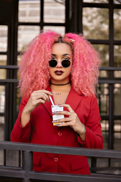 Free Stylish eccentric female with curly pink hair and bright manicure in sunglasses wearing red jacket on blurred background of terrace Stock Photo