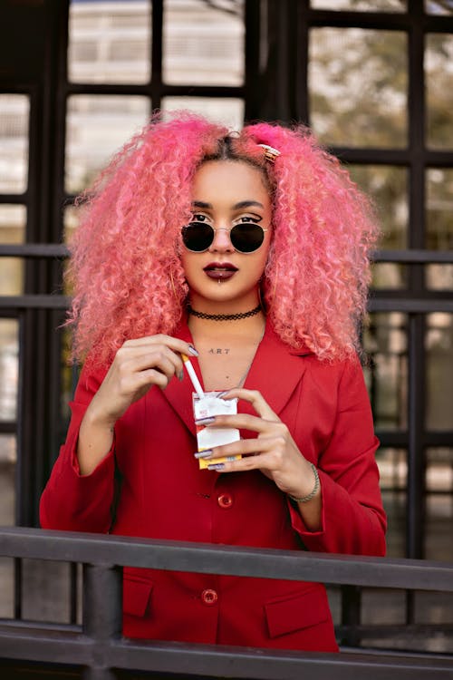 Stylish eccentric female with curly pink hair and bright manicure in sunglasses wearing red jacket on blurred background of terrace