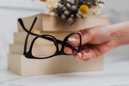 Free A hand holds black eyeglasses near stacked books and dried flowers. Stock Photo
