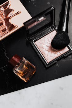 Stylish flat lay of makeup products including highlighter, brush, and perfume bottle on marble surface.