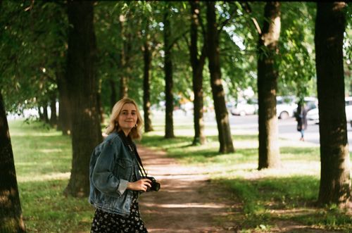 Free Happy young female photographer with camera wearing denim jacket walking through park on sunny day Stock Photo