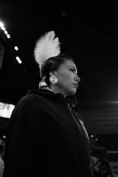 Black and white side view of female in earrings wearing headdress with white feather standing on street with glowing lamps