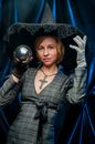 Young woman wearing witch clothes demonstrating small crystal ball while standing against blue background and looking at camera