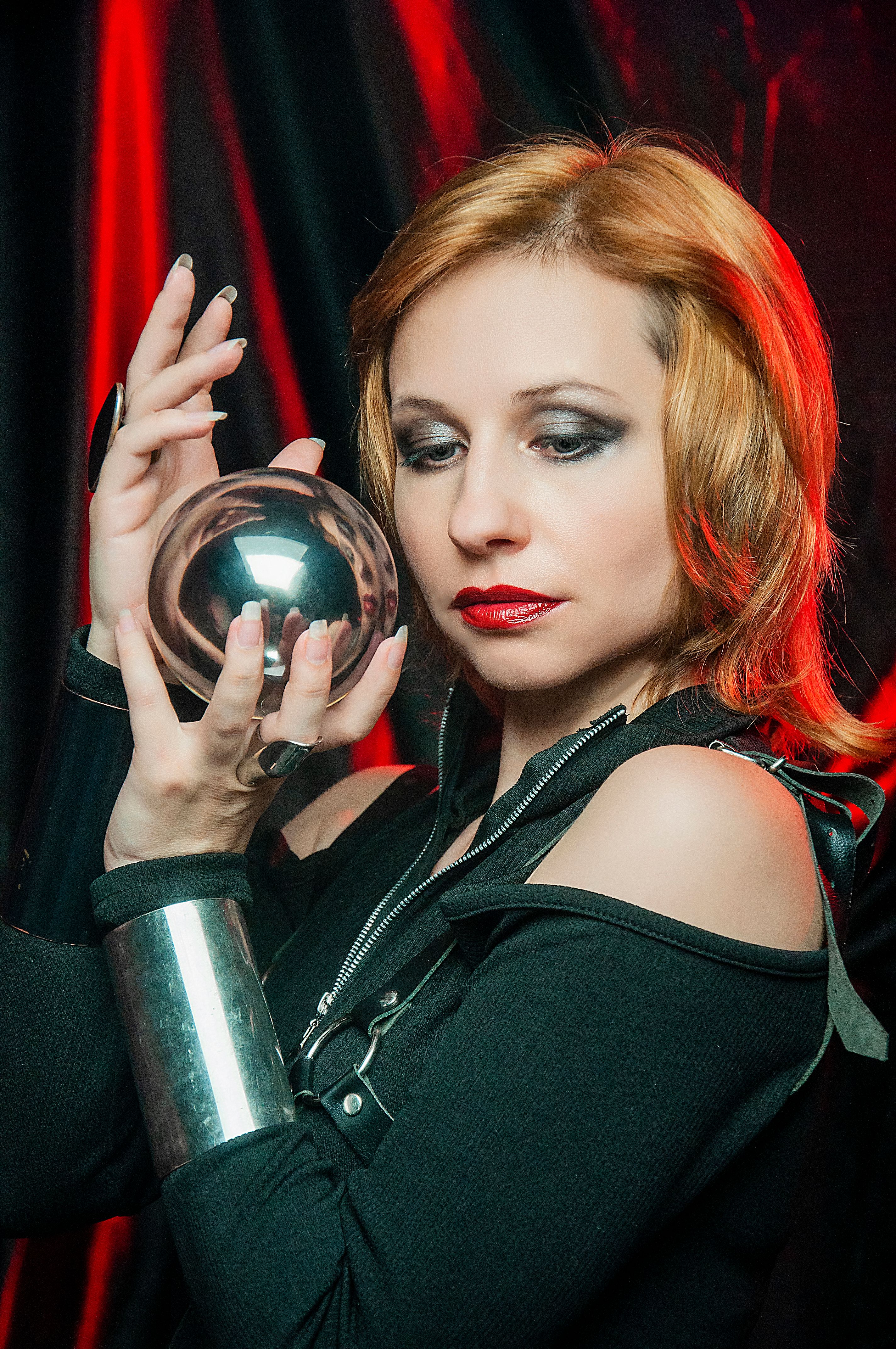 Young lady wearing black outfit with metal bracelets showing shiny ball with reflections while standing in red mystic light