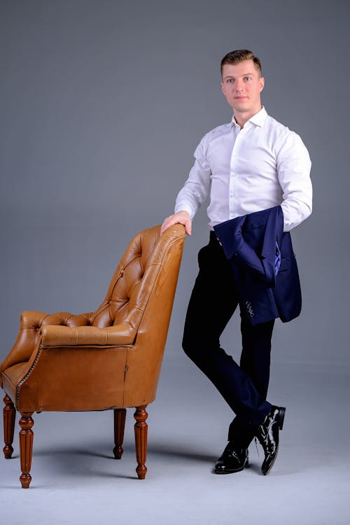 Free Full body of young well dressed male in formal outfit standing in gray studio near comfortable leather armchair Stock Photo