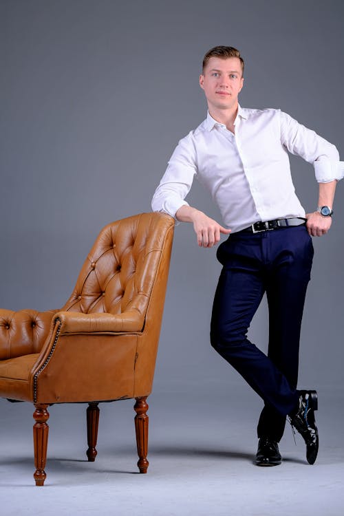 Content young male model in white shirt leaning on vintage leather armchair and looking at camera against gray background