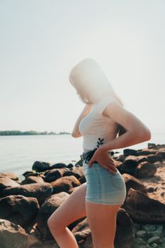 Young woman in denim shorts posing by a sunlit, rocky beach shore, showcasing summer fashion vibes.
