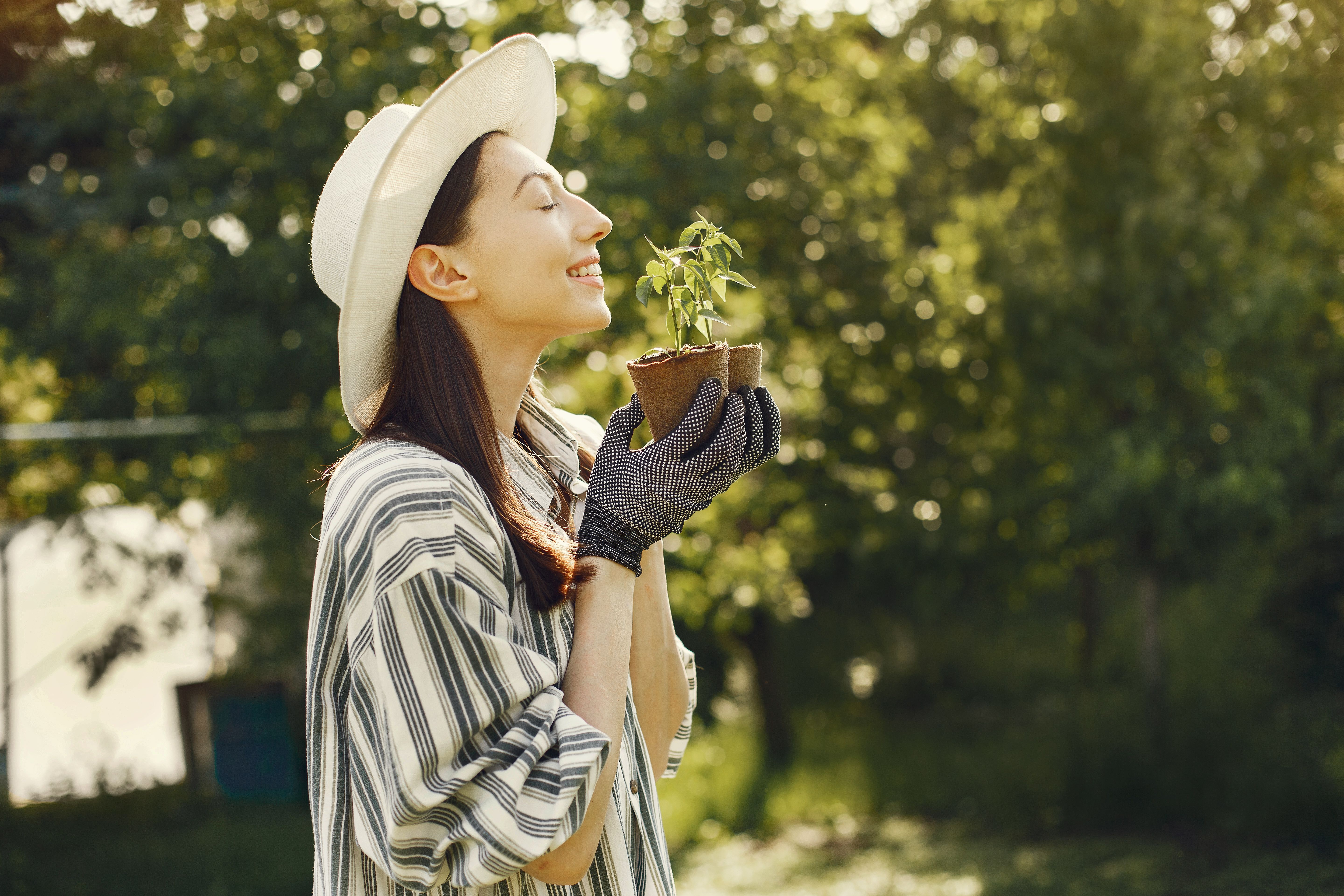 Smiling woman in striped shirt and hat holding a potted plant in a sunlit garden.