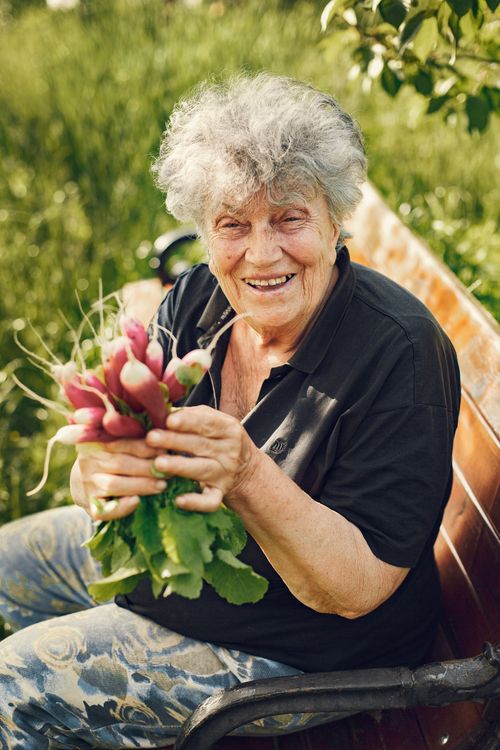 Free Smiling elderly woman sitting outdoors with a bunch of fresh radishes, enjoying a sunny day. Stock Photo