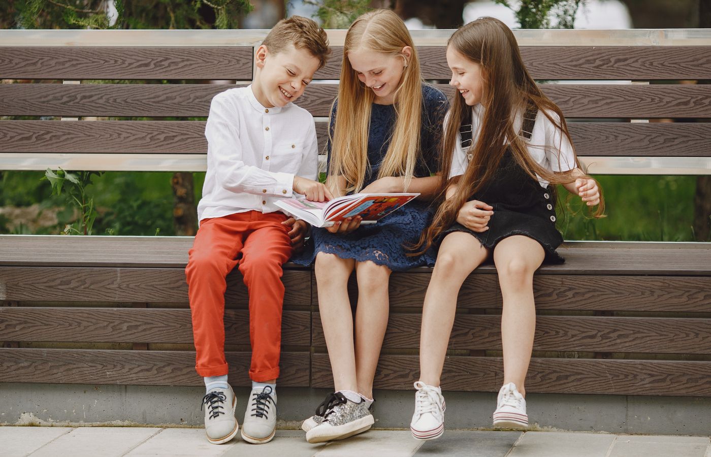 Three happy children reading and laughing together on a park bench outdoors.