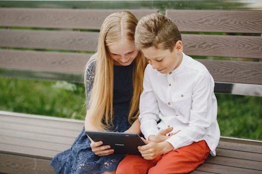 Two kids sitting outdoors, smiling while watching content on a tablet.