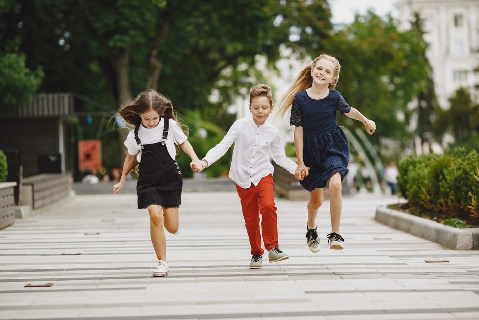 Three children running happily through an urban park, holding hands and smiling.