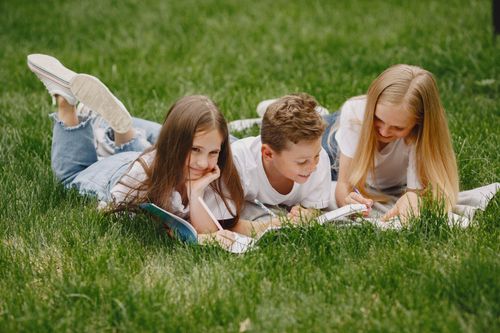 Free Three children lying on grass reading and smiling outdoors. Stock Photo