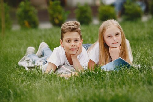 Free Two children lying on grass, reading and enjoying outdoor learning in a sunny spring setting. Stock Photo