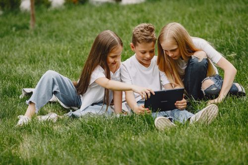 Free Three kids happily sharing a tablet while sitting on lush green grass, enjoying a sunny day. Stock Photo