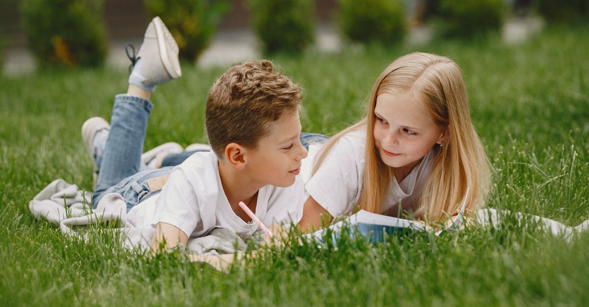 Two children lying on grass, reading and smiling, enjoying a sunny day outdoors.