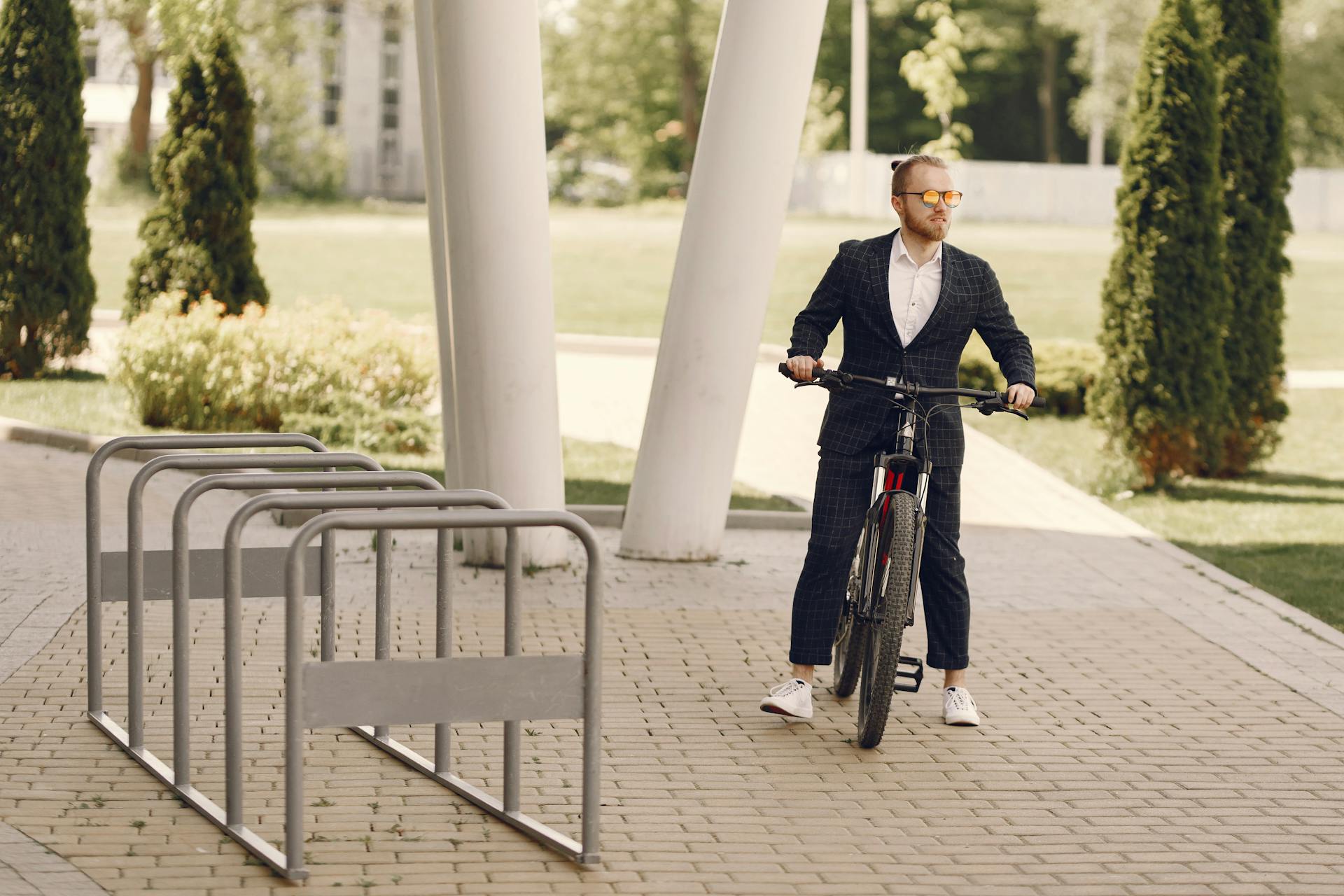 A man in a suit stands with his bicycle at a bike rack in a city park, embracing a healthy lifestyle.