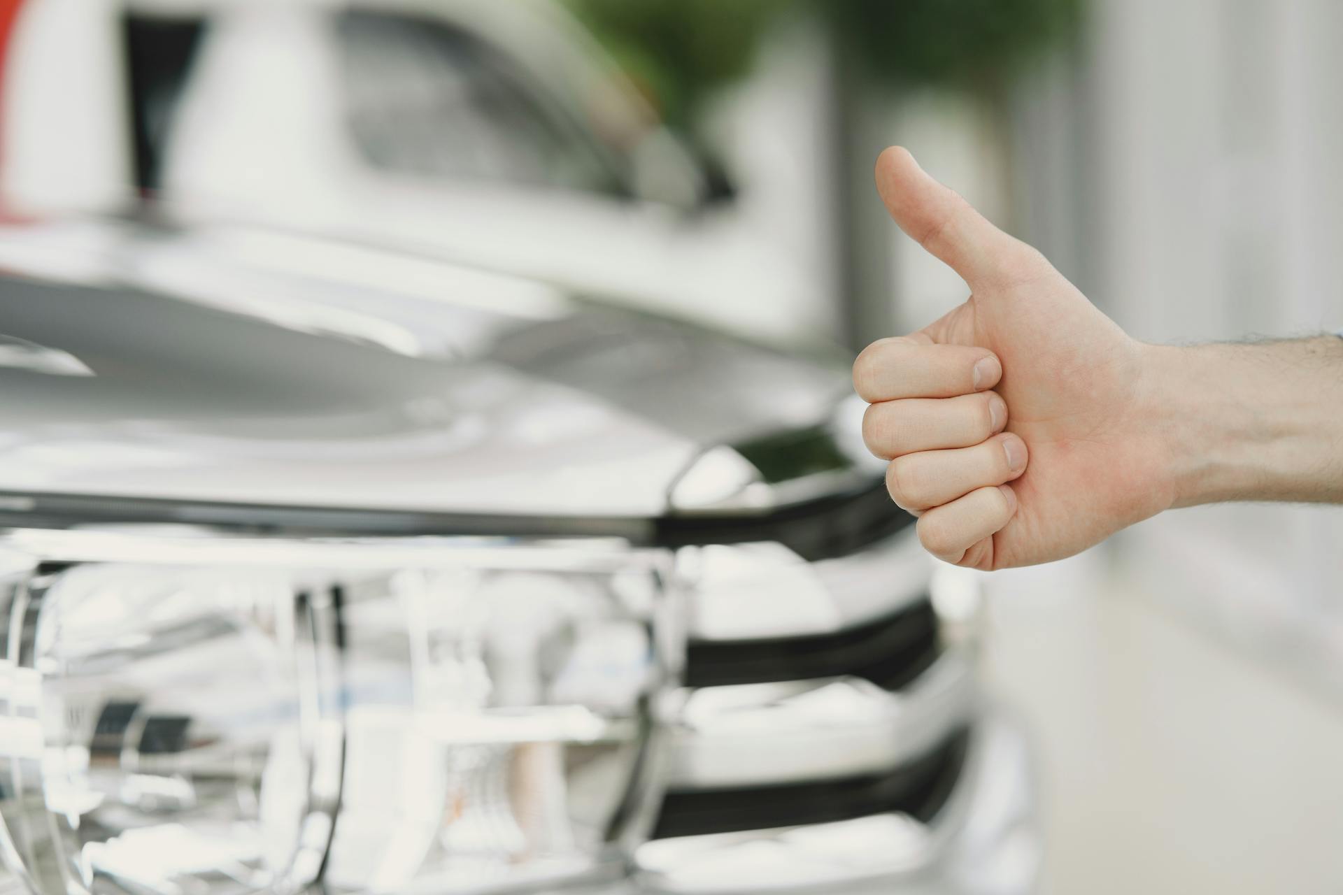 Close-up of a thumbs up gesture next to a car headlight, symbolizing approval or satisfaction.