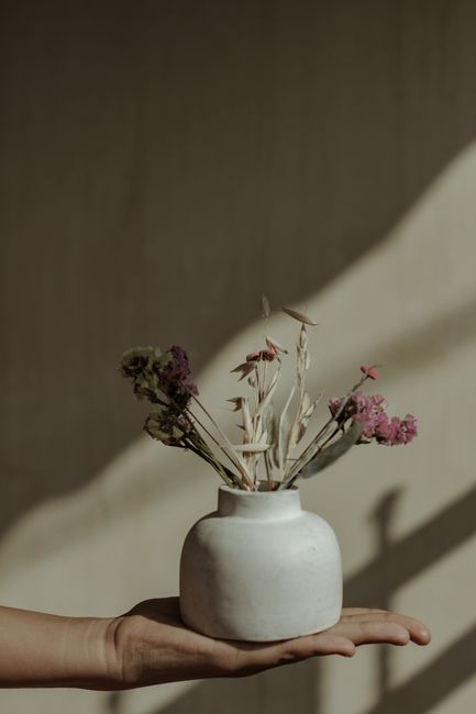 A hand gracefully holds a ceramic vase with dried flowers against a warm backdrop.