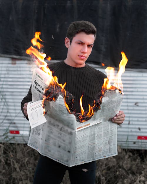 Young male looling at camera while standing near metal fence with burning paper in hands in nature in daylight oitside