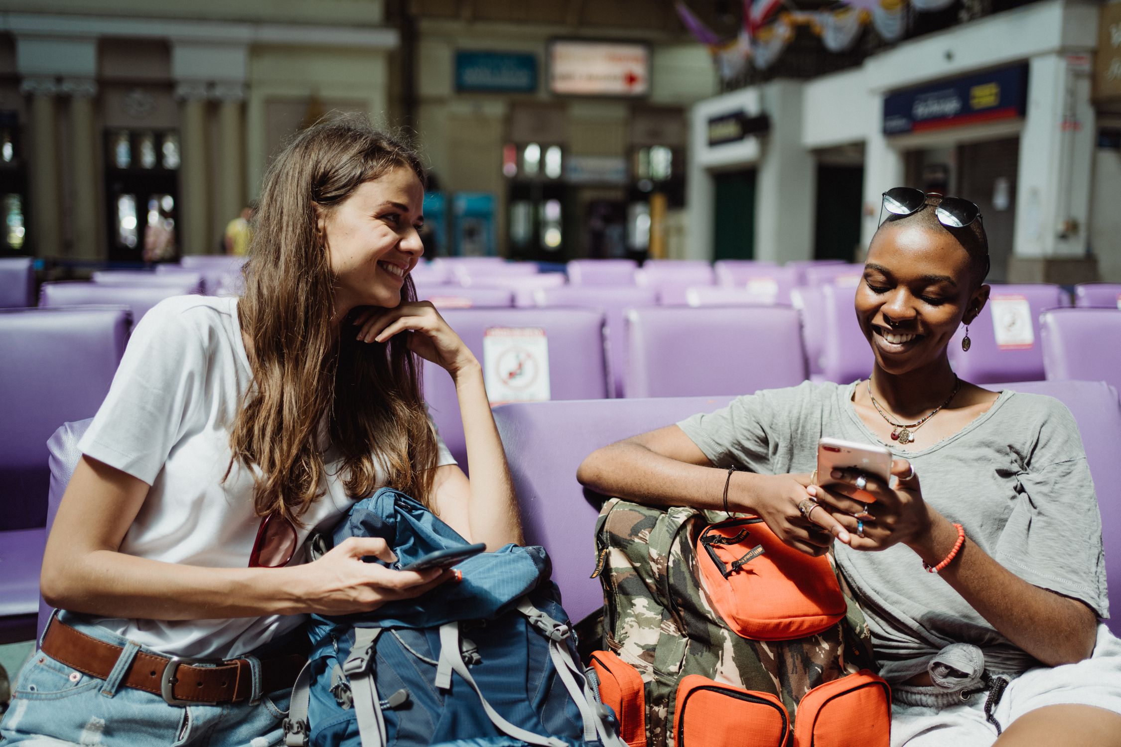Two women travelers smiling with backpacks in a transit waiting area ready for departure