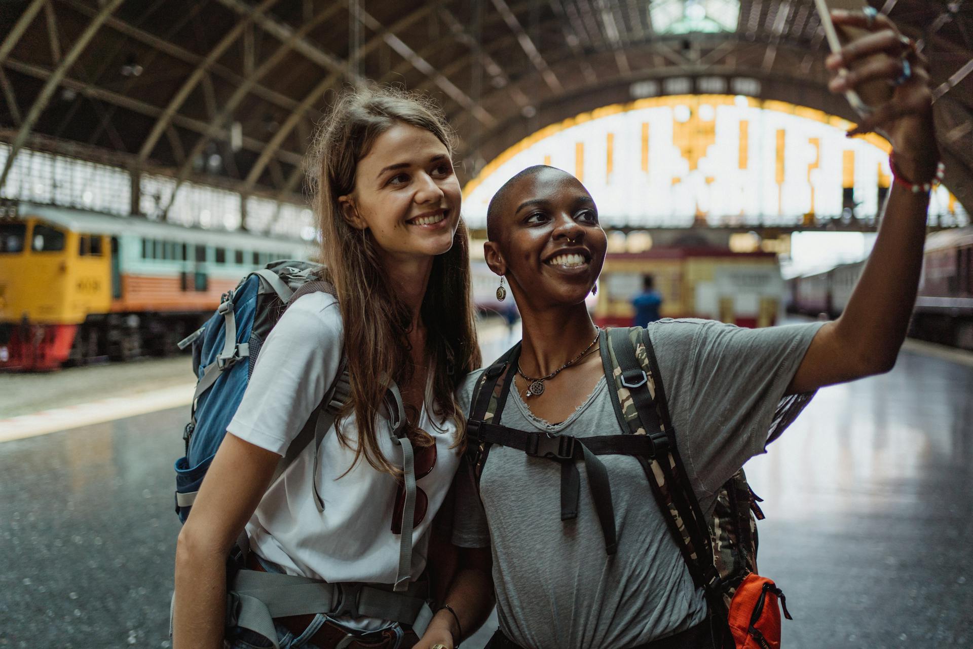 Two women travelers with backpacks taking a selfie at a train station, smiling vividly, capturing their adventures.