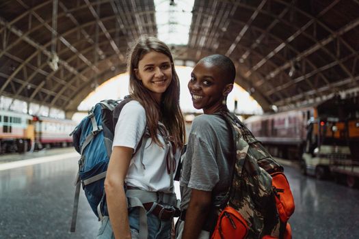 Two women with backpacks smiling at a train station, ready for their travel adventure.