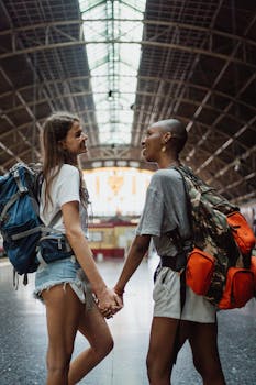 Two women holding hands with backpacks at a train station, expressing love.