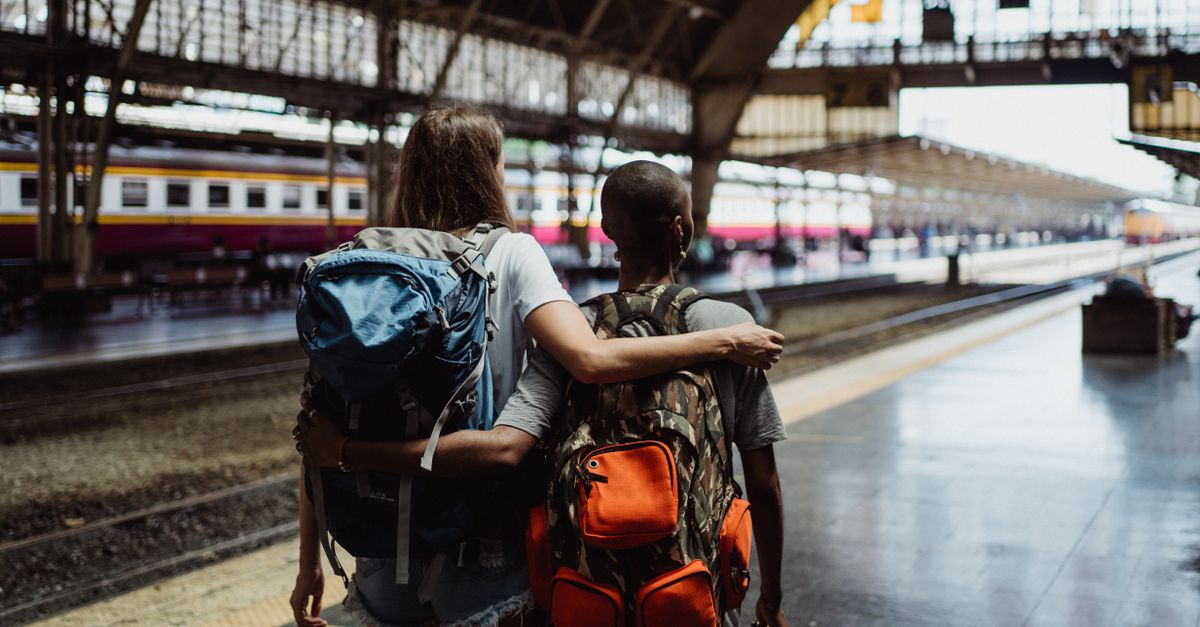 A group of young travelers laughing together on a train platform, backpacks on, sunset in the background