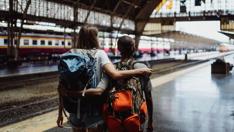 Backpackers waiting at a train station, embracing with arm around shoulders.
