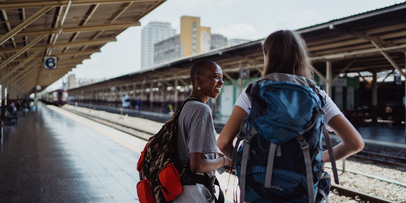 Two diverse female backpackers wait on a sunny railway platform, ready for adventure.